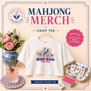Puede incluir: Una camiseta corta blanca con un gráfico de tres fichas de mahjong, incluyendo una con una ilustración de un pájaro, y el texto "BIRD BAM, cheers". La prenda se muestra junto a un jarrón floral azul y blanco, una bebida rosa y fichas.