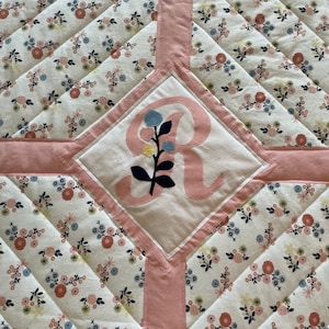 May include: A pink and white quilted baby blanket with a floral pattern and a large embroidered letter "R" in the center.