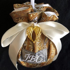 May include: A gold and white fabric ornament with a white satin ribbon and a gold angel wing charm. The ornament is personalized with the initials "JBC".