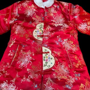 May include: A red, long-sleeved coat with a floral pattern and white fur collar. The coat features embroidered accents and frog closures. The fabric has a shiny, satin-like appearance, with gold and colorful floral details.
