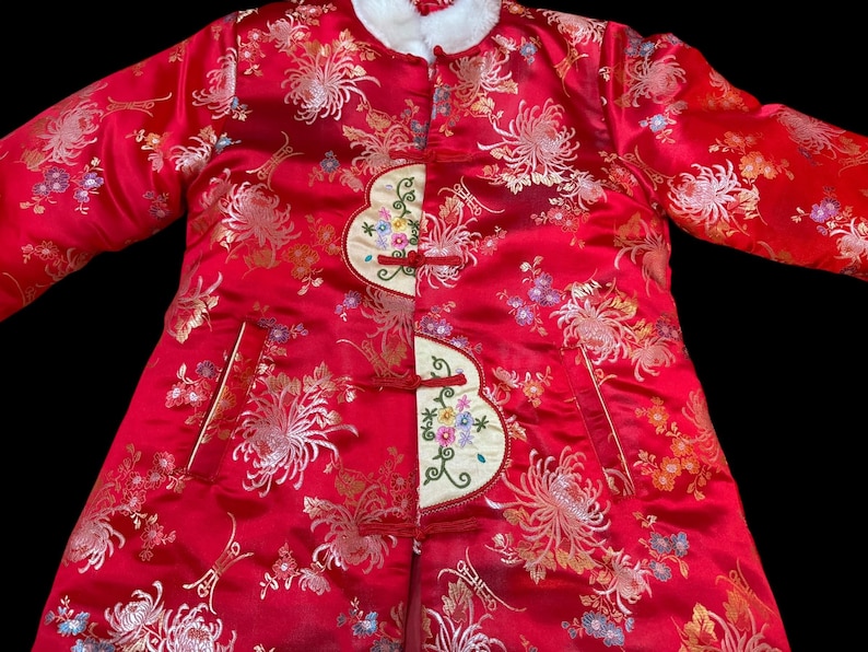 May include: A red, long-sleeved coat with a floral pattern and white fur collar. The coat features embroidered accents and frog closures. The fabric has a shiny, satin-like appearance, with gold and colorful floral details.