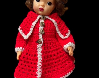 Könnte beinhalten: Eine Vintage-Puppe mit braunem Lockenhaar und blauen Augen, die einen roten gehäkelten Mantel mit weißer Bordüre und goldenen Knöpfen trägt. Die Puppe hat auch eine passende rote gehäkelte Hose. Eine rote Schleife befindet sich im Haar der Puppe.