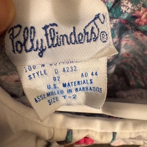 Puede incluir: Primer plano de una etiqueta de tela blanca con el texto azul "Polly Flinders". La etiqueta tambi&eacute;n incluye el texto "100% algod&oacute;n", "STYLE D 4232", "U.S. MATERIALS", "ASSEMBLED IN BARBADOS" y "SIZE T-2".