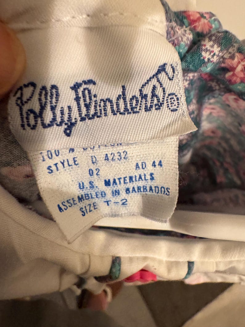 Puede incluir: Primer plano de una etiqueta de tela blanca con el texto azul "Polly Flinders". La etiqueta tambi&eacute;n incluye el texto "100% algod&oacute;n", "STYLE D 4232", "U.S. MATERIALS", "ASSEMBLED IN BARBADOS" y "SIZE T-2".