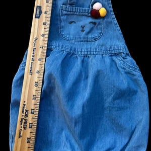May include: Blue denim baby overalls with a bear face design on the chest pocket. The overalls have adjustable straps and a decorative pom-pom accent. The ruler shows the overalls are approximately 16 inches long.