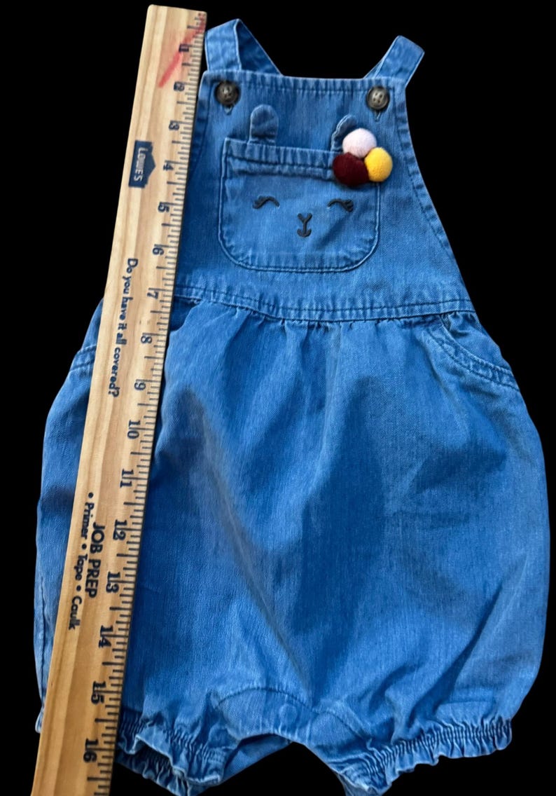 May include: Blue denim baby overalls with a bear face design on the chest pocket. The overalls have adjustable straps and a decorative pom-pom accent. The ruler shows the overalls are approximately 16 inches long.