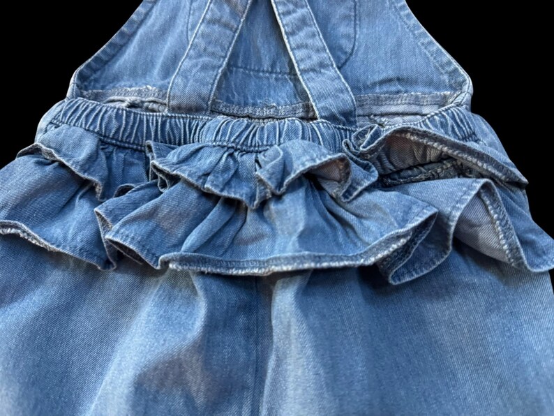 May include: Close-up of a light blue denim overall dress. The dress features ruffled detailing around the waist and a gathered elastic waistband. The straps cross in the back. The denim has a classic, faded wash.