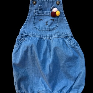 May include: Blue denim baby romper with a bunny face design on the front pocket. The romper features adjustable straps, button closures, and a decorative pom-pom accent. The garment is designed for infants and toddlers.
