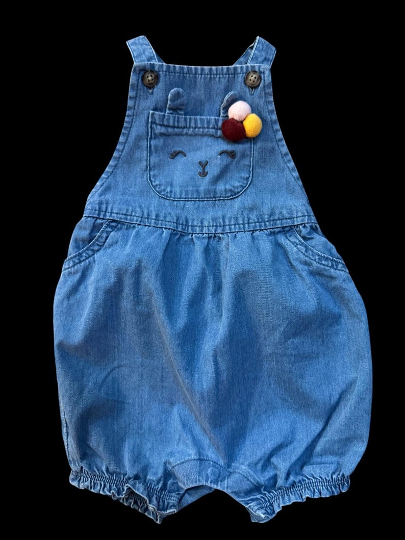 May include: Blue denim baby romper with a bunny face design on the front pocket. The romper features adjustable straps, button closures, and a decorative pom-pom accent. The garment is designed for infants and toddlers.