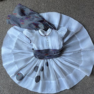 Pode incluir: Um vestido de tule branco com uma faixa xadrez preta e branca e listras coloridas. O vestido tem um corpete branco com decote redondo e mangas curtas. A saia é em camadas e tem uma bainha festonada. Há quatro bolinhas na saia.