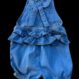 May include: A light blue denim romper with ruffled detailing. The romper features criss-cross straps in the back and elasticized leg openings. The waistline has a layered ruffle design, adding a playful touch to the garment.