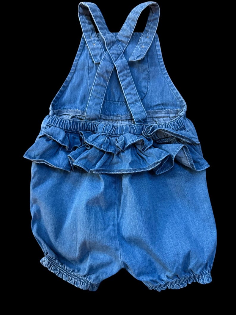 May include: A light blue denim romper with ruffled detailing. The romper features criss-cross straps in the back and elasticized leg openings. The waistline has a layered ruffle design, adding a playful touch to the garment.