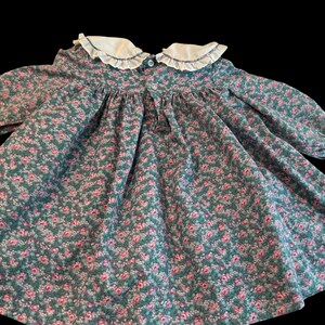 Puede incluir: Vestido vintage con estampado floral. Presenta un cuello blanco con volantes y mangas largas. La tela es verde azulado con un estampado de peque&ntilde;as flores rosas.
