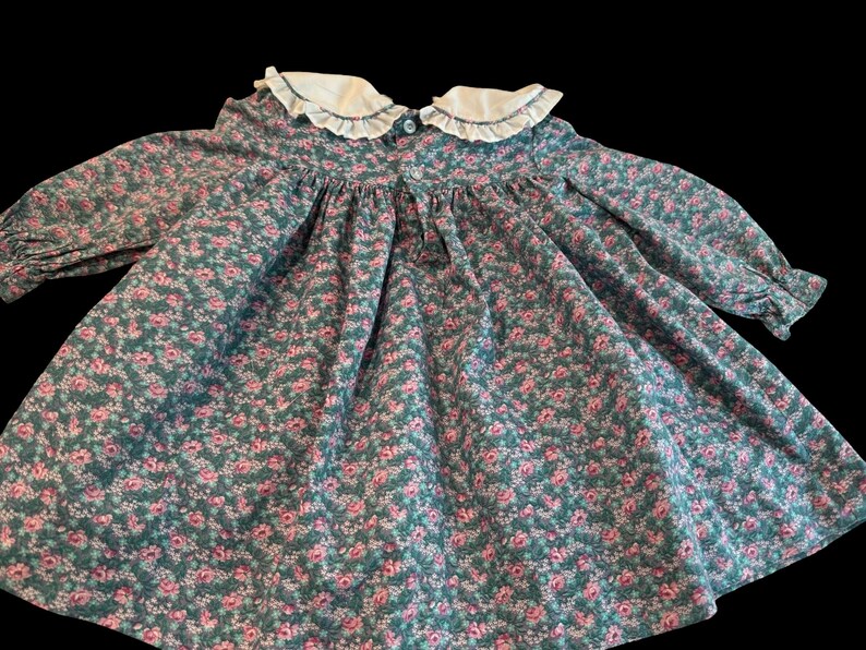 Puede incluir: Vestido vintage con estampado floral. Presenta un cuello blanco con volantes y mangas largas. La tela es verde azulado con un estampado de peque&ntilde;as flores rosas.