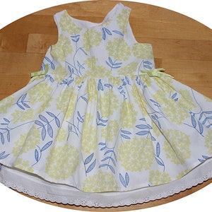 Puede incluir: Un vestido blanco sin mangas con estampado floral amarillo y azul. El vestido tiene una falda fruncida y un ribete de encaje blanco.