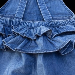 May include: Close-up of a blue denim overall with ruffled detailing. The overall features adjustable straps and an elastic waistband. The ruffled skirt adds a playful touch to the garment. The denim fabric has a classic, medium wash.