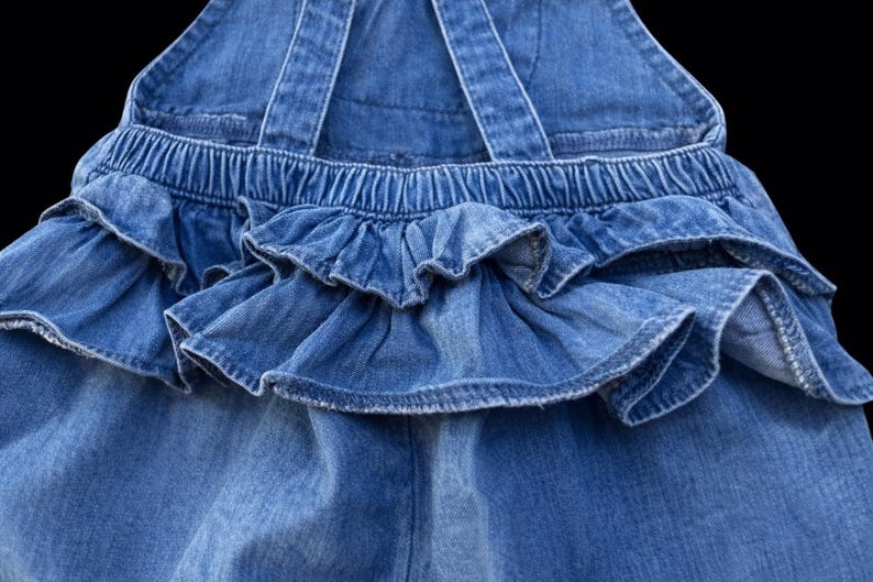 May include: Close-up of a blue denim overall with ruffled detailing. The overall features adjustable straps and an elastic waistband. The ruffled skirt adds a playful touch to the garment. The denim fabric has a classic, medium wash.