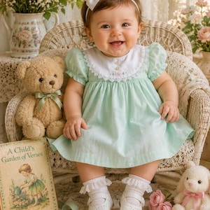 May include: A smiling baby wearing a mint green dress with a white collar and lace trim, seated in a wicker chair. The baby wears a white bow headband, white socks with lace trim, and white shoes. A teddy bear, a book titled "A Child's Garden of Verses", and a stuffed bunny are nearby.