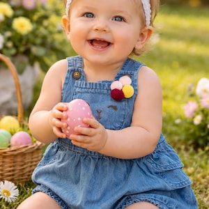 May include: A smiling baby wearing a denim romper and a white bow headband, holding a pink Easter egg with white polka dots. A basket of colorful eggs and flowers are in the background.