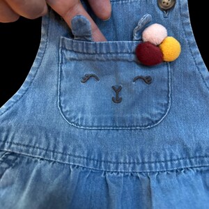 May include: Blue denim overall dress with a pocket featuring an embroidered bunny face. The pocket is adorned with a cluster of colorful pom-poms. The dress has a button closure on the strap.