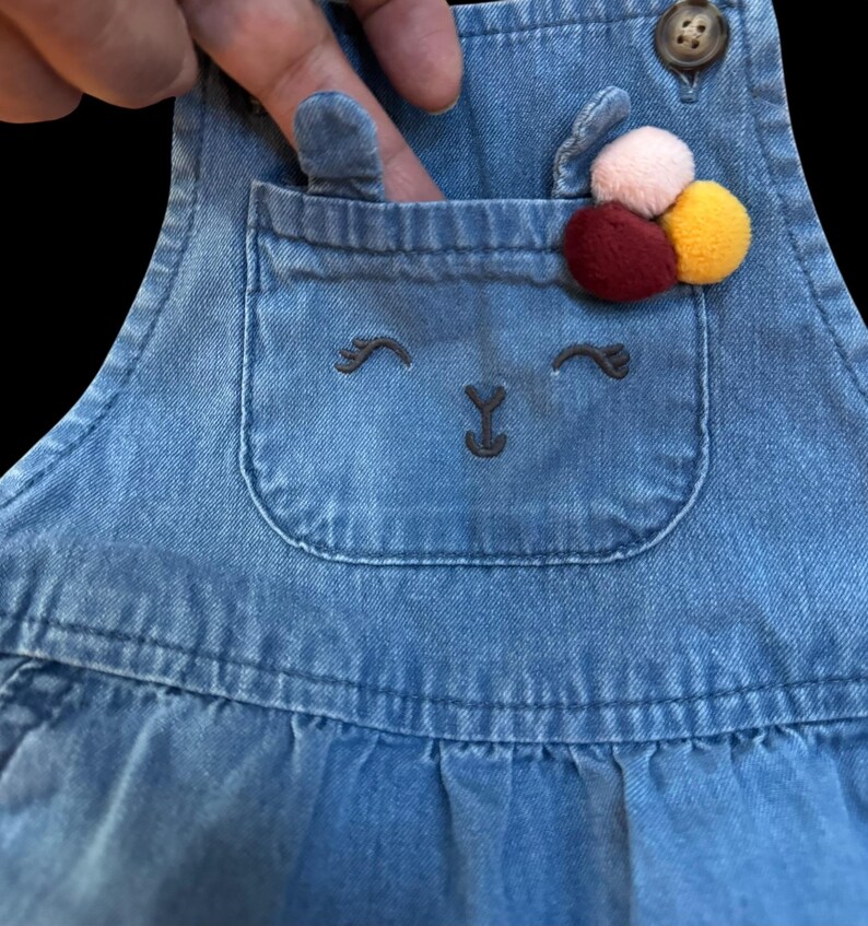 May include: Blue denim overall dress with a pocket featuring an embroidered bunny face. The pocket is adorned with a cluster of colorful pom-poms. The dress has a button closure on the strap.