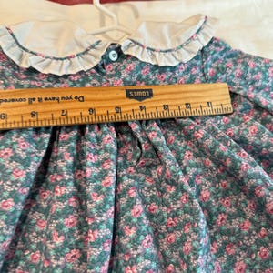 Puede incluir: Un vestido vintage con estampado floral, cuello blanco con volantes y cierre de botones. El vestido presenta un fondo verde azulado con estampados florales rosas y blancos. Una regla de madera est&aacute; colocada sobre el vestido, midiendo unos 25 cm.