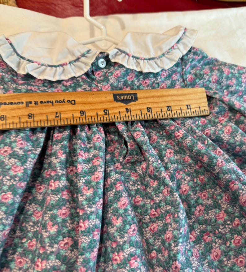 Puede incluir: Un vestido vintage con estampado floral, cuello blanco con volantes y cierre de botones. El vestido presenta un fondo verde azulado con estampados florales rosas y blancos. Una regla de madera est&aacute; colocada sobre el vestido, midiendo unos 25 cm.
