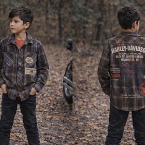 Może przedstawiać: Młoda osoba w brązowo-niebiesko-pomarańczowej koszuli w kratę Harley-Davidson i ciemnych dżinsach. Koszula ma naszywki i tekst z przodu i z tyłu. Tło to rozmyta scena na zewnątrz.