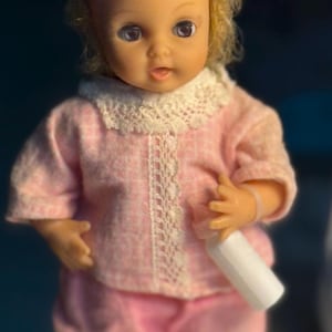 Könnte beinhalten: Eine Vintage-Puppe mit blonden Haaren, die ein rosa-weißes Kleid mit Spitzenkragen trägt. Die Puppe hält eine weiße Flasche in ihrer rechten Hand.