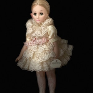 May include: A porcelain doll with blonde hair, green eyes, and a white lace dress with a pink ribbon. The doll is wearing white tights and white shoes. The doll has a tag that says "Just Dottie" and "September 1990".