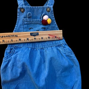 May include: Blue denim baby overalls with a bib pocket and decorative pom-poms. The overalls have adjustable straps and button closures. The bib features a smiling face design. A wooden ruler is placed across the overalls.