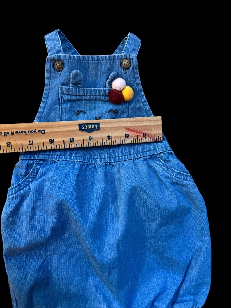 May include: Blue denim baby overalls with a bib pocket and decorative pom-poms. The overalls have adjustable straps and button closures. The bib features a smiling face design. A wooden ruler is placed across the overalls.