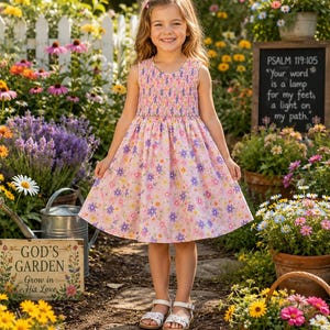 May include: A young girl wearing a pink floral dress with a smocked bodice and white sandals stands in a garden. The dress features a pattern of purple and yellow flowers. A sign reads "GOD'S GARDEN Grow in His Love."