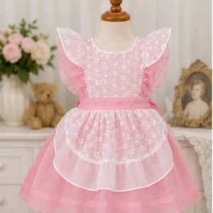 May include: A pink dress featuring a white floral embroidered overlay on the bodice and a matching white floral apron-style panel at the waist. The dress has ruffled sleeves, a round neckline, and a full skirt, displayed on a mannequin.