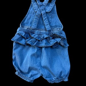 May include: Blue denim romper with ruffled detailing. The romper features adjustable straps that cross in the back and elasticized leg openings. The garment is designed for a baby or toddler.