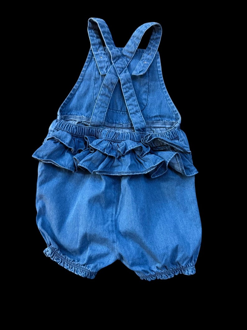 May include: Blue denim romper with ruffled detailing. The romper features adjustable straps that cross in the back and elasticized leg openings. The garment is designed for a baby or toddler.