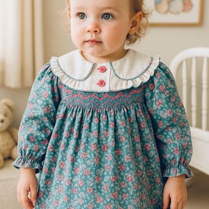 Puede incluir: Vestido turquesa con estampado floral, cuello blanco y botones rosas. El vestido tiene mangas largas con pu&ntilde;os con volantes y un corpi&ntilde;o smock. El vestido est&aacute; dise&ntilde;ado para un beb&eacute; o un ni&ntilde;o peque&ntilde;o.