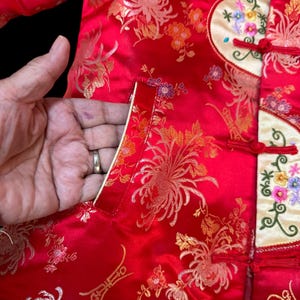 May include: Red satin jacket with gold floral embroidery. The jacket features a mandarin collar, frog closures, and a cream-colored panel with colorful floral embroidery. The pocket is lined with cream-colored satin.
