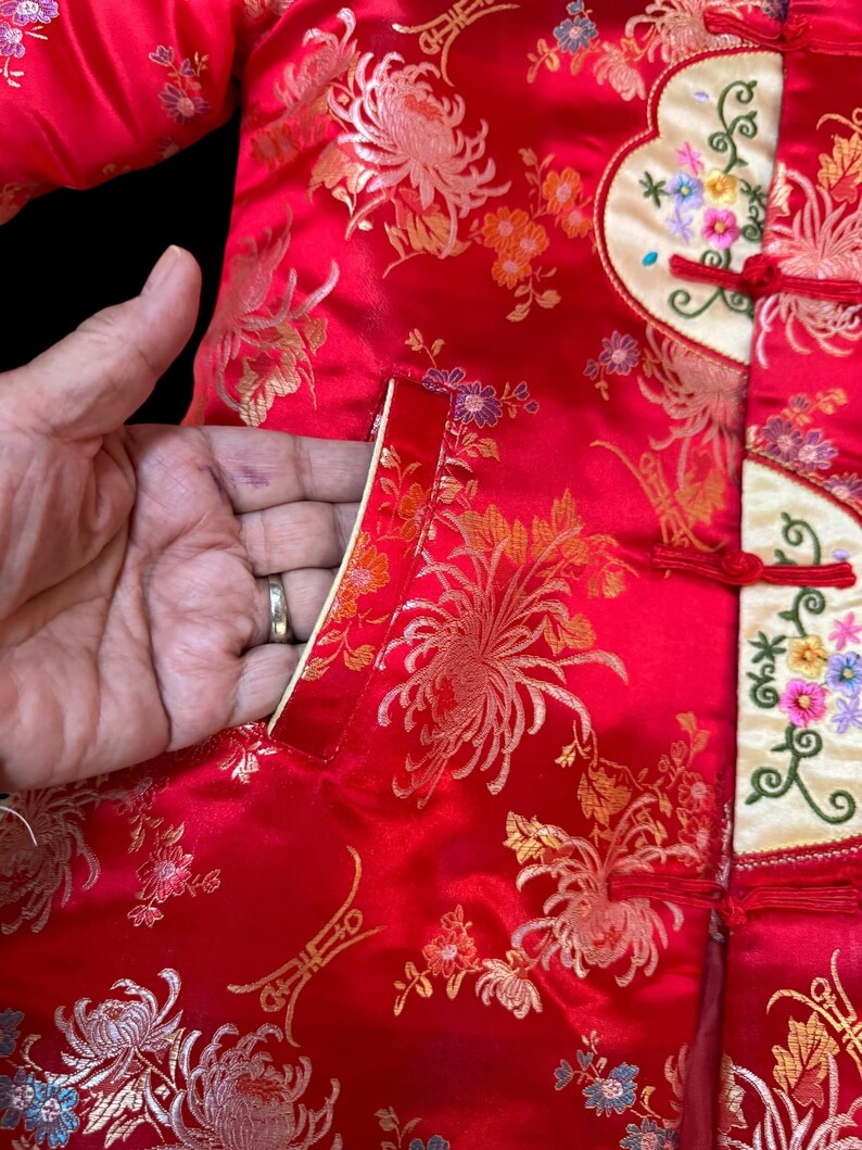 May include: Red satin jacket with gold floral embroidery. The jacket features a mandarin collar, frog closures, and a cream-colored panel with colorful floral embroidery. The pocket is lined with cream-colored satin.