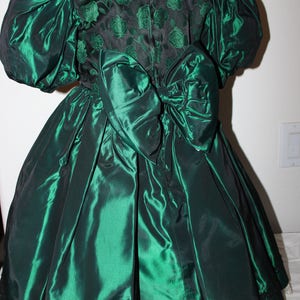 Puede incluir: Un vestido verde oscuro con un corpiño de patrón de rosas negras, mangas abullonadas y un lazo grande en la cintura. La falda es de tela verde oscuro lisa con un borde negro en el dobladillo y una capa de encaje blanco visible debajo.