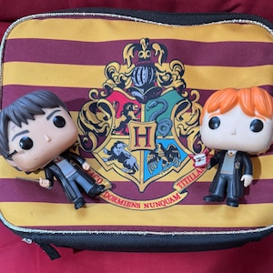 May include: A black lunch bag with a gold and burgundy striped pattern and a Hogwarts crest. Two Funko Pop figures of Harry Potter and Ron Weasley are in front of the bag. Harry is holding a wand, and Ron is holding a small object.