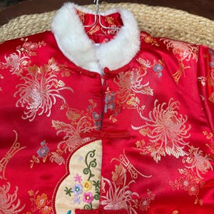 May include: A red satin jacket with a white faux fur collar. The jacket features an embroidered floral design in gold, orange, and blue. It has red frog closures and a cream-colored embroidered panel with colorful flowers.