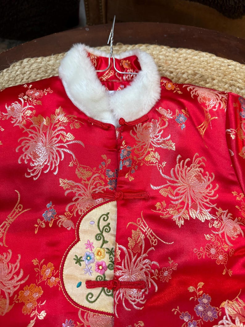 May include: A red satin jacket with a white faux fur collar. The jacket features an embroidered floral design in gold, orange, and blue. It has red frog closures and a cream-colored embroidered panel with colorful flowers.
