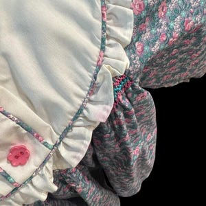 Puede incluir: Un vestido de estilo vintage con un corpi&ntilde;o blanco y mangas florales. El corpi&ntilde;o tiene un bot&oacute;n de flor rosa y bordes con volantes. Las mangas presentan un estampado floral rosa y verde. El vestido tiene un aspecto suave y texturizado.