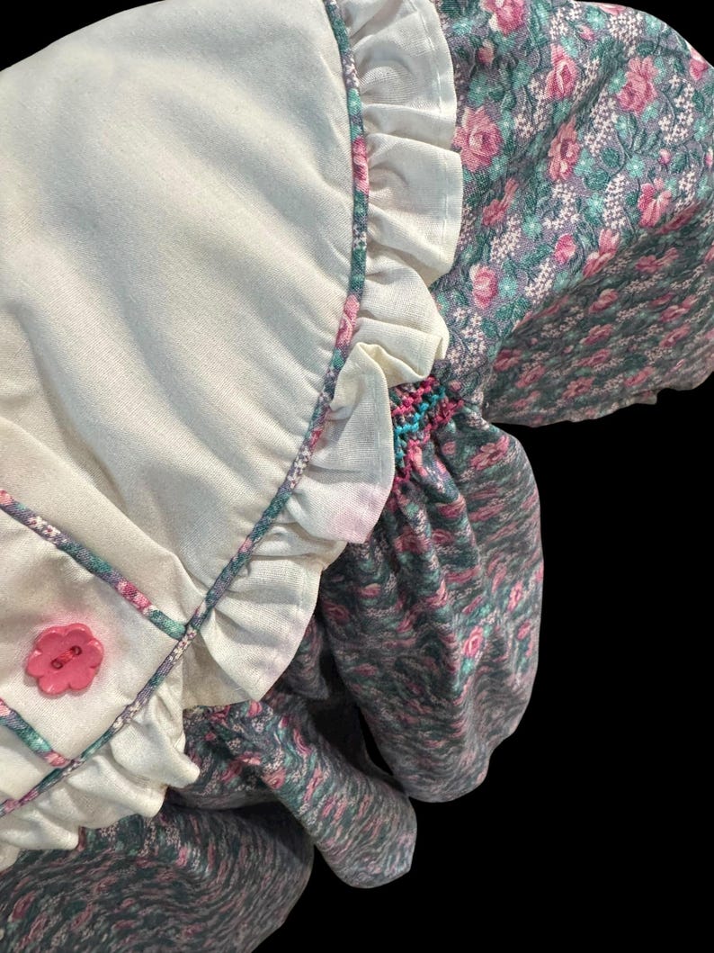 Puede incluir: Un vestido de estilo vintage con un corpi&ntilde;o blanco y mangas florales. El corpi&ntilde;o tiene un bot&oacute;n de flor rosa y bordes con volantes. Las mangas presentan un estampado floral rosa y verde. El vestido tiene un aspecto suave y texturizado.