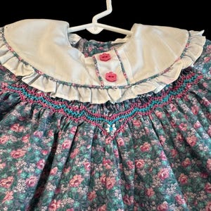 Puede incluir: Vestido con estampado floral con cuello blanco y botones rosas. El vestido presenta un corpi&ntilde;o fruncido con costuras turquesas y rosas. La tela tiene un estampado de rosas rosas y hojas verdes.