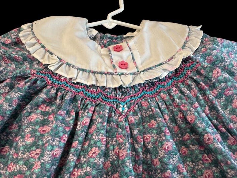 Puede incluir: Vestido con estampado floral con cuello blanco y botones rosas. El vestido presenta un corpi&ntilde;o fruncido con costuras turquesas y rosas. La tela tiene un estampado de rosas rosas y hojas verdes.