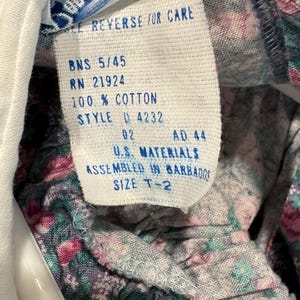 Puede incluir: Primer plano de una etiqueta de ropa con el texto "SEE REVERSE FOR CARE", "100% COTTON" y "SIZE T-2". La etiqueta es blanca con texto negro. La tela alrededor de la etiqueta tiene un estampado floral con flores rosas y verdes.