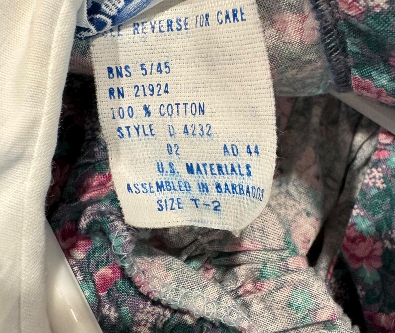 Puede incluir: Primer plano de una etiqueta de ropa con el texto "SEE REVERSE FOR CARE", "100% COTTON" y "SIZE T-2". La etiqueta es blanca con texto negro. La tela alrededor de la etiqueta tiene un estampado floral con flores rosas y verdes.