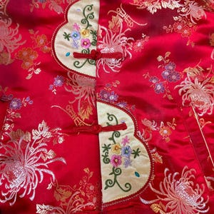May include: A red satin jacket with gold floral embroidery and cream-colored panels. The panels feature colorful floral embroidery and red frog closures. The jacket's design is reminiscent of traditional Asian garments.